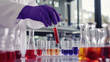 © Саша Федюк - A scientist in a lab coat and purple gloves carefully adds red liquid from a test tube into a series of small glass containers filled with colorful solutions on a laboratory bench.
