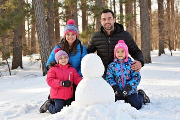 Group portrait of cheerful family sitting in snow around small snowman in winter forest and looking at camera with smiles enjoying vacation in nature