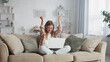 © MIKHAIL - Young woman sitting comfortably on a sofa, laptop resting on her lap, raising her arms in joyful celebration of success while working from home in a cozy living room setting