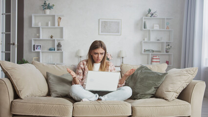 Young woman sitting with crossed legs on a beige sofa in her living room, looking confused and gesturing in frustration while using a laptop with a blank white screen
