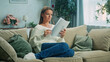 © MIKHAIL - Young woman sitting comfortably on a sofa, reading documents in a cozy living room, enjoying peaceful moments at home while surrounded by natural light and relaxing decor