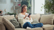 © MIKHAIL - Young woman wearing casual clothes sitting comfortably on a sofa in her living room, using her smartphone and enjoying a relaxing day at home