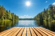 © Halina Berah - The tranquil beauty of a Finnish summer day is captured by the shimmering lake, an old wooden pier, and the sun’s rays filtering through the blue sky above the forest