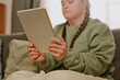 © AnnaStills - Young woman with Down syndrome sitting in living room, holding digital tablet, reading book online