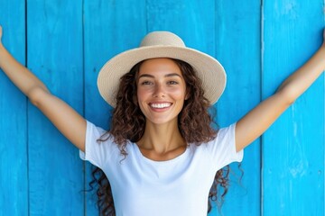 Wall Mural - A joyful woman wearing a straw hat stands against a bright blue wooden background, smiling with her arms raised, radiating happiness and summer vibes.