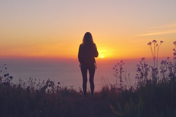 Poster - Woman silhouetted against a vibrant sunset overlooking the ocean, wildflowers in the foreground.