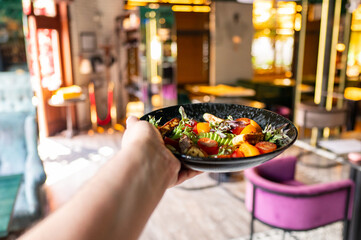 Wall Mural - A hand holds a plate of vibrant salad, featuring colorful vegetables, against a stylishly decorated restaurant interior with soft lighting and modern furnishings.