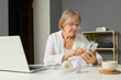 © sementsova321 - A senior woman in a white shirt checks her smartphone at a home office desk. She is focused, with a laptop nearby and a bowl and headphones on the table
