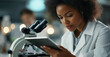 © GustavsMD - Focused female scientist in a lab coat examines data on a tablet while working with a microscope in a modern laboratory