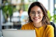© Vilaysack - A smiling woman in glasses sits at a laptop in a bright, modern workspace, surrounded by greenery and vibrant decor.