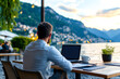 © F-STUDIO - A person works on a laptop at a scenic lakeside setting, surrounded by mountains and greenery under a beautiful sky.