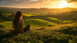 © roei - A woman sitting on a grassy hill overlooking rolling vineyards in a remote valley in New Zealand, Oceania with lush greenery and a golden sunset