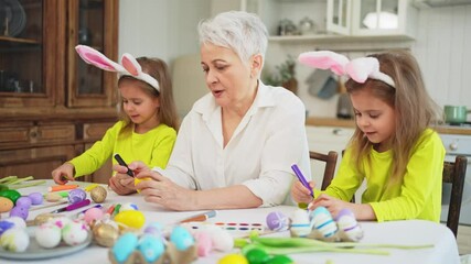 Wall Mural - Happy Easter. Two little girls sisters twins grandmother painting eggs enjoying time together. Happy family grandma granddaughters child kids preparing for Easter. Spring Christian festival tradition