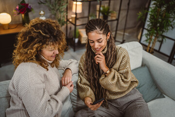 two adult female friends sit on sofa and enjoy while use cellphone