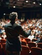 © vefimov - A man in a suit giving a speech with his hands outstretched, standing on stage as an audience listens intently.