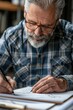 © vefimov - Elderly individual working on architectural designs, wearing glasses and plaid shirt.