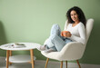 © Pixel-Shot - Young African-American woman with cup of tea sitting in white armchair near green wall