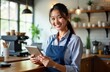 © Maryna - Asian female cafe owner smiles happily using tablet near counter. Modern cafe setting. Successful small business owner. Friendly, confident barista. Digital tech for cafe management. Charming cafe