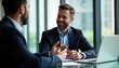 © Maryna - Two businesspeople in suits communicate indoors. Sitting at desk in office, appear to discussion. Man on right smiling, gestures. Laptop sits on desk in front of. Likely colleagues business partners.