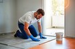 © Maryna - Handyman installs gray tiles on floor inside apartment. Focused on work, wearing blue work gloves. Bucket of mortar sits nearby. Room light walls, open doorway. Renovation improvement in progress.