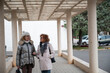 © C&A - Middle aged women walking outdoors under a covered walkway, wearing winter clothes, engaging in a casual conversation in a serene suburban environment