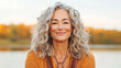 © VERTEX SPACE - woman with curly gray hair smiles warmly, wearing orange sweater, against serene outdoor backdrop with autumn colors. Her joyful expression radiates positivity