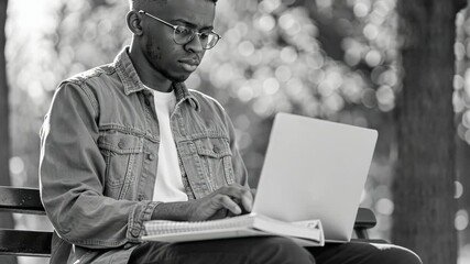 Wall Mural - Young man wearing glasses sits on a bench with a laptop open in front of him. He is typing on the laptop while looking at a book. Concept of focus and productivity as the man works on his laptop