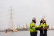 © kamonrat - Two engineers in safety helmets and high-visibility jackets discuss work while holding a tablet and clipboard at a power plant site,with transmission towers and industrial structures in the background