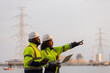 © kamonrat - Two engineers in safety helmets and high-visibility jackets discuss work while holding a tablet and clipboard at a power plant site,with transmission towers and industrial structures in the background