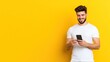 © julio - Young man smiling while using a smartphone against a yellow background.