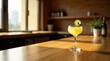 © Mali - A refreshing yellow cocktail garnished with a lime wedge and mint, sits on a sunlit wooden table in a modern setting.