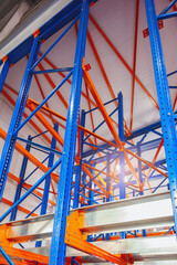 Fototapete - A tall, blue and orange rack system is situated in a large warehouse. The rack system is made up of multiple levels, with each level holding various items
