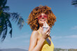 © SHOTPRIME STUDIO - Young woman enjoying ice cream under the sunny sky with palm trees in the background Her curly hair and bright outfit add to the summer vibe