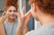 © Cavan Images - Young woman applying moisturizing cream on face looking in mirror