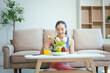 © NanSan - A happy Asian woman in sportswear sits on a sofa at a table, enjoying a salad bowl, fruits, fresh vegetables, orange juice, and energy supplements after a home fitness workout