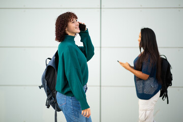 Wall Mural - Two women using mobile phones while walking with backpacks, the background is a plain wall