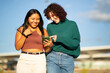 © Paula - Two young women smiling while looking at a phone outdoors