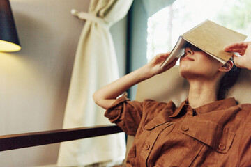 Canvas Print - A relaxed woman enjoying a moment of calm, covering her eyes with a book, dressed in a cozy brown outfit, conveying serenity in a warm, softly lit room