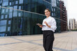 © muse studio - Modern professional woman using a tablet outside a contemporary office building in an urban setting