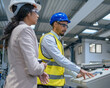 © 24K-Production - Indian female factory boss training African American male worker, in blue helmet and yellow vest, to work on manufacturing machinery.