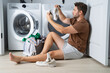 © Panacea Stock  - A man is sitting on the floor with his feet up and holding a pair of dirty socks. He is surrounded by a pile of clothes and a basket. Concept of laziness and disorganization