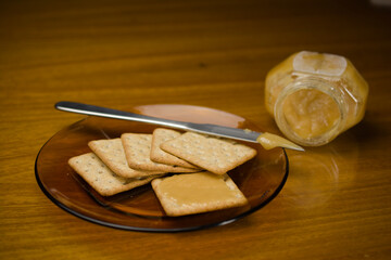 Wall Mural - A plate of crackers topped with golden honey, accompanied by a jar of honey and a knife on a wooden table, offering a simple and sweet snack option.