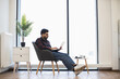 © sofiko14 - Young Indian man sitting on chair using laptop in bright modern living room. Relaxed work from home setup with casual clothing and natural lighting.