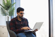 © sofiko14 - Young Indian male in casual attire using laptop seated by large window. Bright natural light fills modern workspace, creating an inspiring environment for remote work.