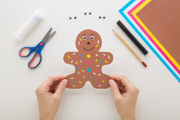 Wall Mural - Young adult woman hands holding brown gingerbread man shape from application paper on light gray table background. Making decoration elements. Closeup. Flat lay. Point of view shot. Top down view.