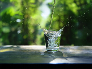 Poster - Water pours into a glass, creating a splash against a lush green, blurred background.  Sunlight reflects on the surface.