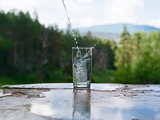 Aufkleber - Clear water pours into a glass on a rustic surface, with a lush green forest backdrop.  The scene evokes refreshment and nature.