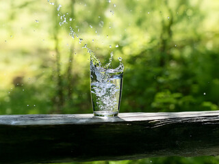 Aufkleber - A glass of water sits on a rustic wooden surface,  water splashing dramatically, set against a vibrant green, natural backdrop.  The sunlight creates glistening droplets.