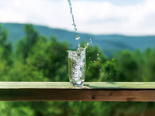 Aufkleber - Crystal clear water pours into a glass, creating a splash.  The glass sits on a wooden surface with a lush green mountain backdrop.  Refreshing, idyllic summer scene.