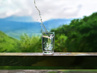 Aufkleber - Clear water splashes into a glass on a wooden surface overlooking a lush, mountainous landscape.  The scene evokes freshness and tranquility.